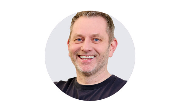 Porträt von Sebastian Hünermann, Hünermann Physiotherapie, breit lächelnd mit kurzen Haaren, Dreitagebart und schwarzem T-Shirt.
