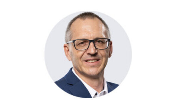 Porträtaufnahme von Sales Manager Dirk Geitner mit kurzer Frisur, Brille und dunklem Sakko vor neutralem, hellem Hintergrund.