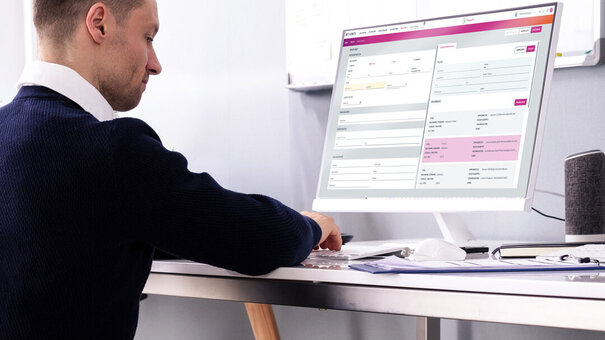 Seitliche Aufnahme einer männlichen Person am Schreibtisch, die mit Tastatur arbeitet und einen Monitor mit Formularansicht bedient.