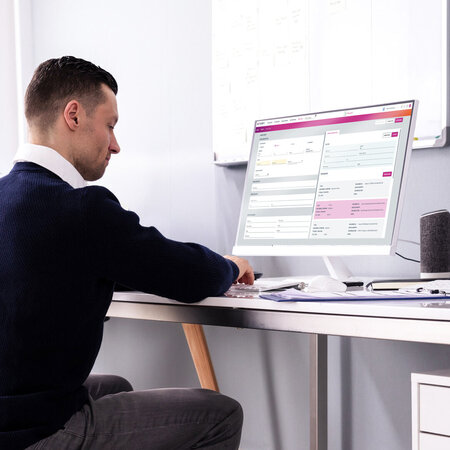 Seitliche Aufnahme einer männlichen Person am Schreibtisch, die mit Tastatur arbeitet und einen Monitor mit Formularansicht bedient.