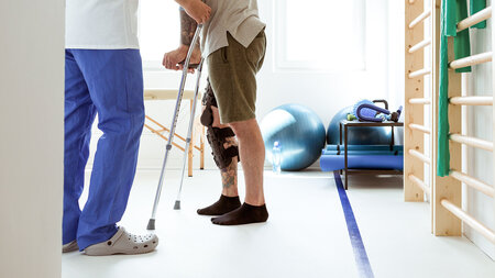 Physiotherapeut unterstützt Patienten mit Knieorthese und Gehhilfe bei Reha-Übungen im Therapieraum – digitale Rehasport-Abrechnung mit NOVENTI.