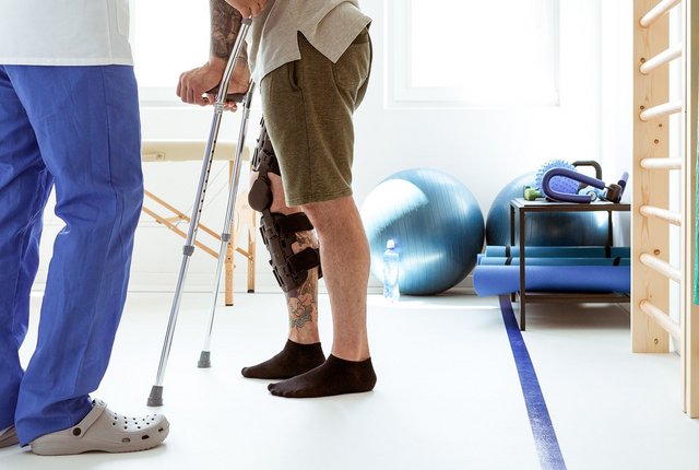 Physiotherapeut unterstützt Patienten mit Knieorthese und Gehhilfe bei Reha-Übungen im Therapieraum – digitale Rehasport-Abrechnung mit NOVENTI.