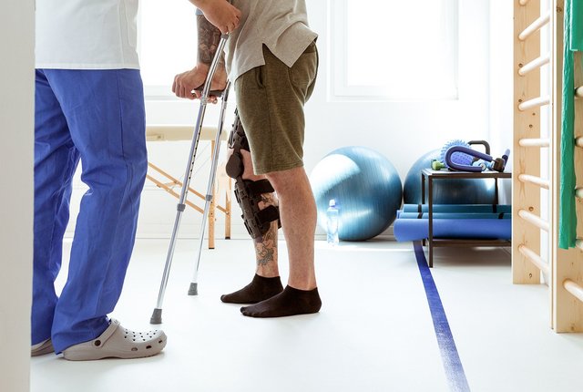 Physiotherapeut unterstützt Patienten mit Knieorthese und Gehhilfe bei Reha-Übungen im Therapieraum – digitale Rehasport-Abrechnung mit NOVENTI.