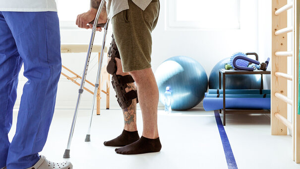Physiotherapeut unterstützt Patienten mit Knieorthese und Gehhilfe bei Reha-Übungen im Therapieraum – digitale Rehasport-Abrechnung mit NOVENTI.