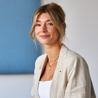 Porträtaufnahme von Marketingmanagerin Lisa Barsig mit hochgesteckten, hellen Haaren und hellem Blazer vor blauem Hintergrund.